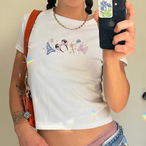 Op de afbeelding: Witte crop top met geborduurde cartoonafbeeldingen, waaronder een tovenaars hoed, Minnie Mouse oren, een toverstaf en een vlinder. De top heeft een ronde hals en korte mouwen.