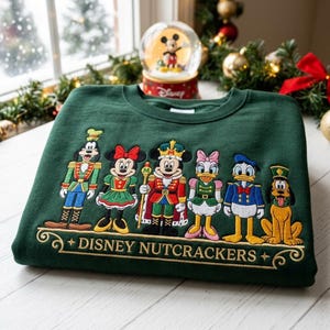 May include: A dark green crewneck sweatshirt featuring an embroidered design of six Disney characters dressed as nutcrackers. The characters include Goofy, Minnie Mouse, Mickey Mouse, Daisy Duck, Donald Duck, and Pluto. The text reads DISNEY NUTCRACKERS.