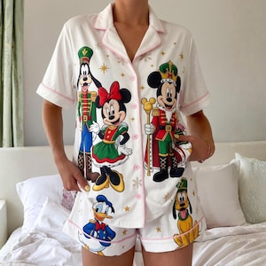 May include: A white short-sleeve button-down pajama set featuring a festive print of Disney characters including Mickey Mouse, Minnie Mouse, Goofy, Donald Duck, and Pluto dressed in holiday attire with gold stars and snowflakes.