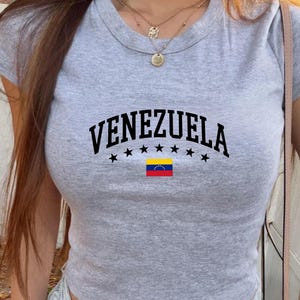 May include: A gray athletic heather crop top with the word "VENEZUELA" in black, arched above a row of black stars and the Venezuelan flag. The shirt has a crew neck and short sleeves.