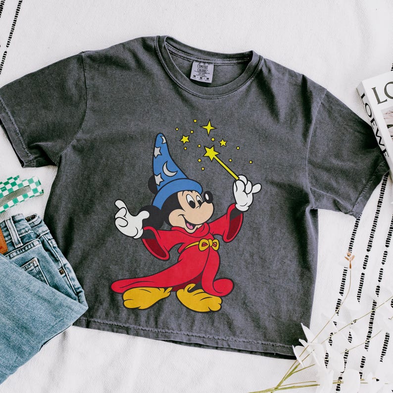 Puede incluir: Camiseta gris oscuro con un dibujo de Mickey Mouse como hechicero, sosteniendo una varita con estrellas. Mickey lleva un sombrero azul con lunas y estrellas, una t&uacute;nica roja y zapatos amarillos. La camiseta se muestra con vaqueros azules.