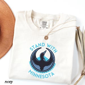 May include: Ivory-colored t-shirt with the text "STAND WITH MINNESOTA" in light blue. The design features a stylized black bird with a red eye inside a blue circle. A gold necklace is draped across the shirt.