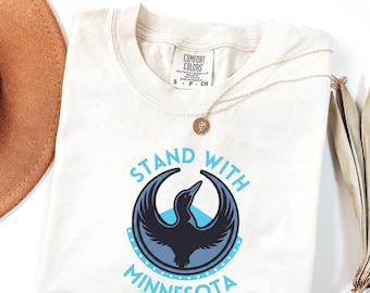 Puede incluir: Camiseta de color marfil con el texto "STAND WITH MINNESOTA" en azul claro. El diseño presenta un pájaro negro estilizado con un ojo rojo dentro de un círculo azul. Un collar dorado está sobre la camiseta.