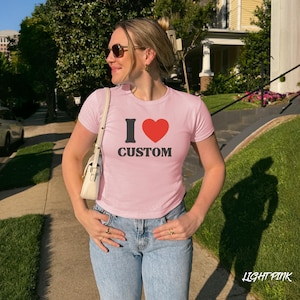 May include: Light pink t-shirt with the text "I &hearts; CUSTOM" in black and red. The shirt is a crew neck style and is paired with blue jeans. The image is taken outdoors in natural light.