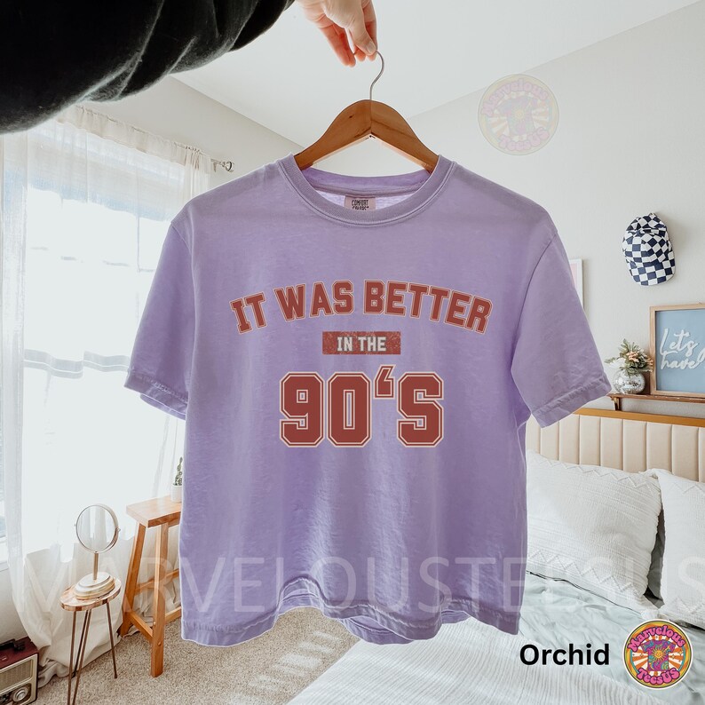 May include: A light purple short-sleeve t-shirt with a crew neckline. The front features the text "IT WAS BETTER IN THE 90'S" printed in a distressed, dark orange font with a white outline. The shirt is displayed on a wooden hanger.