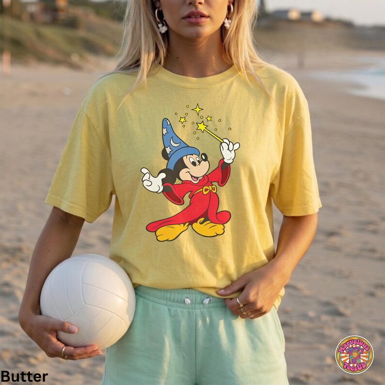 Puede incluir: Camiseta amarilla con un dibujo de Mickey Mouse como hechicero, sosteniendo una varita con estrellas. El personaje de dibujos animados viste una t&uacute;nica roja y un sombrero de mago azul. La camiseta se combina con pantalones cortos verde claro.