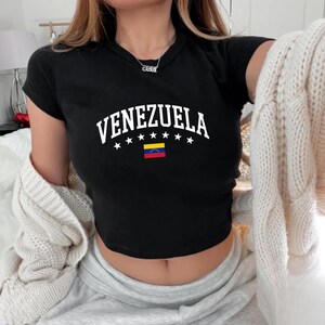 May include: Black crop top with the word "VENEZUELA" in white, arched above a row of white stars and the Venezuelan flag. The shirt has short sleeves and a round neckline.
