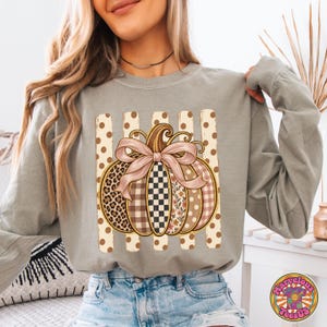 May include: A sage green long-sleeve sweatshirt featuring a central graphic of a pumpkin decorated with a pink bow. The pumpkin has segments with leopard print, checkered, floral, and plaid patterns, set against a background of vertical polka-dot stripes.
