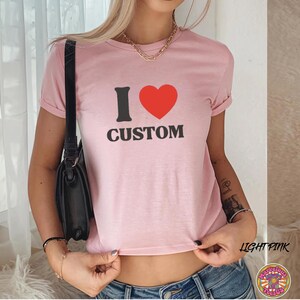 May include: Light pink t-shirt with the text "I &hearts; CUSTOM" in black and red. The shirt has rolled-up sleeves and is made of a soft material. The words "LIGHT PINK" are written in a script font on the bottom right.