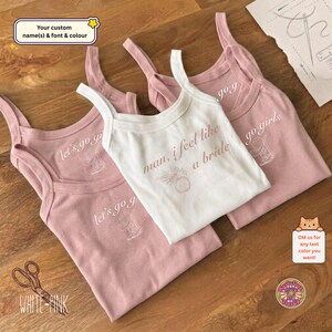 May include: Three sleeveless tank tops in pink and white cotton. Two pink tops feature the white text let's go girls with a cowboy boot graphic. One white top features the brown text man, i feel like a bride with a diamond ring graphic.