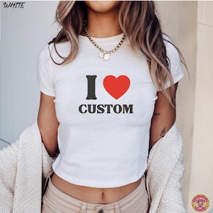 May include: White crop top with the text "I &hearts; CUSTOM". The "I" is black, the heart is red, and the word "CUSTOM" is black. The top is short-sleeved and has a crew neck.