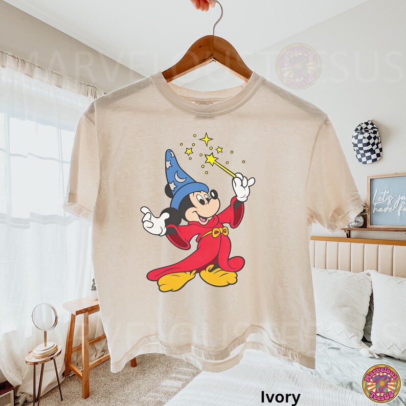 Puede incluir: Camiseta de color marfil con un dibujo de Mickey Mouse como hechicero, sosteniendo una varita con estrellas. Mickey lleva un sombrero azul con estrellas, una t&uacute;nica roja y zapatos amarillos. La camiseta est&aacute; colgada en una percha de madera.