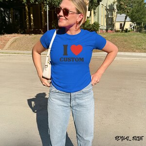 May include: Royal blue t-shirt with the text "I ❤️ CUSTOM" in black and red. The shirt is short-sleeved and has a crew neck. The model is wearing light blue jeans.