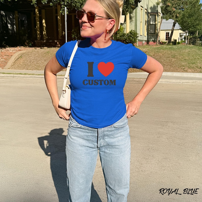May include: Royal blue t-shirt with the text "I ❤️ CUSTOM" in black and red. The shirt is short-sleeved and has a crew neck. The model is wearing light blue jeans.