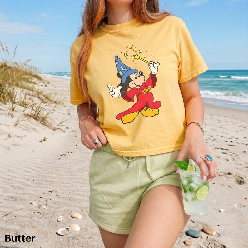 Puede incluir: Camiseta amarilla con un dise&ntilde;o de Mickey Mouse de dibujos animados como mago, sosteniendo una varita con estrellas. La camiseta se combina con pantalones cortos verde claro. La palabra "Butter" est&aacute; impresa en la parte inferior izquierda de la imagen.