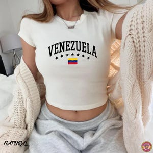 May include: Cream-colored crop top with the word "VENEZUELA" in black, arched above a row of black stars and the Venezuelan flag. The top has a scoop neck and short sleeves. The word "NATURAL" is printed in the bottom left corner.