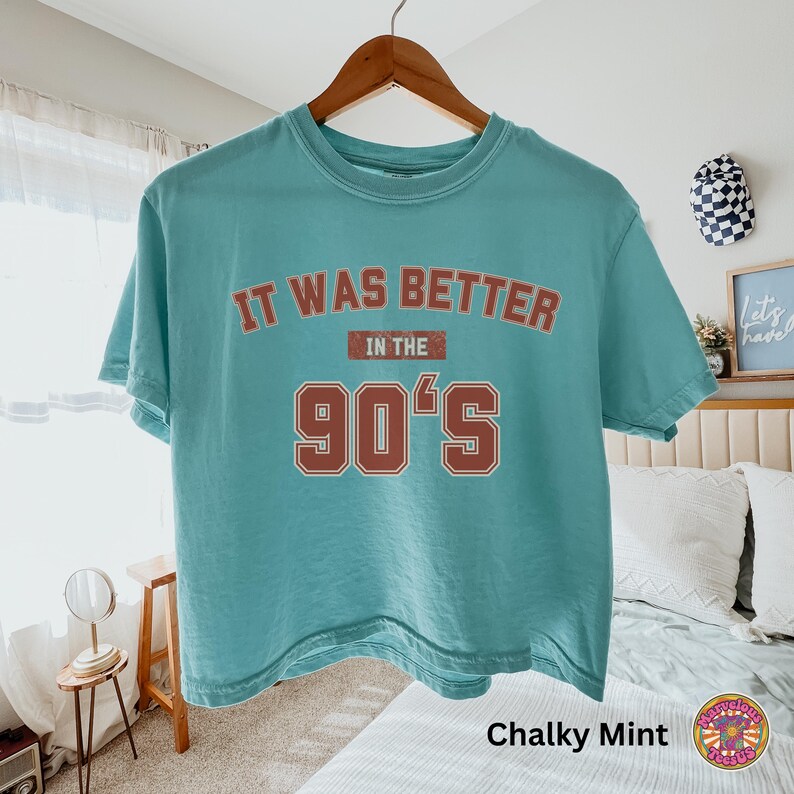 May include: A chalky mint green short-sleeve t-shirt featuring the text "IT WAS BETTER IN THE 90'S" printed in a dark red collegiate-style font with a white outline. The shirt is displayed on a wooden hanger against a neutral indoor setting.