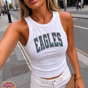 May include: A white ribbed tank top featuring the word EAGLES printed in dark blue with a thin yellow outline across the chest. The garment has a high crew neckline and sleeveless cut, paired with white denim shorts and a tan clutch bag.