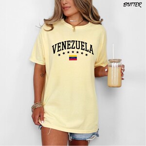 May include: A light yellow t-shirt with the word "VENEZUELA" in black, arched above a row of black stars and the Venezuelan flag. The shirt is short-sleeved and has a relaxed fit. The word "BUTTER" is printed in the upper right corner.