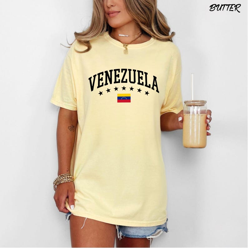 May include: A light yellow t-shirt with the word "VENEZUELA" in black, arched above a row of black stars and the Venezuelan flag. The shirt is short-sleeved and has a relaxed fit. The word "BUTTER" is printed in the upper right corner.