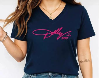 May include: A navy blue V-neck t-shirt featuring the word Dolly in a stylized pink script font, followed by the text xoxo and two small pink hearts. The shirt has short sleeves and a simple, clean design across the front chest area.