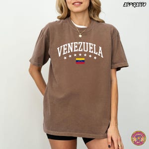 May include: Espresso-colored t-shirt with the word "VENEZUELA" in white arched text, with white stars above and a Venezuelan flag graphic below. The shirt is short-sleeved and has a crew neck.