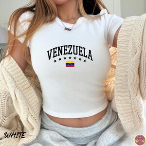 May include: White crop top with the word "VENEZUELA" in black, arched above a row of black stars and a small Venezuelan flag. The shirt is short-sleeved and has a scoop neck.