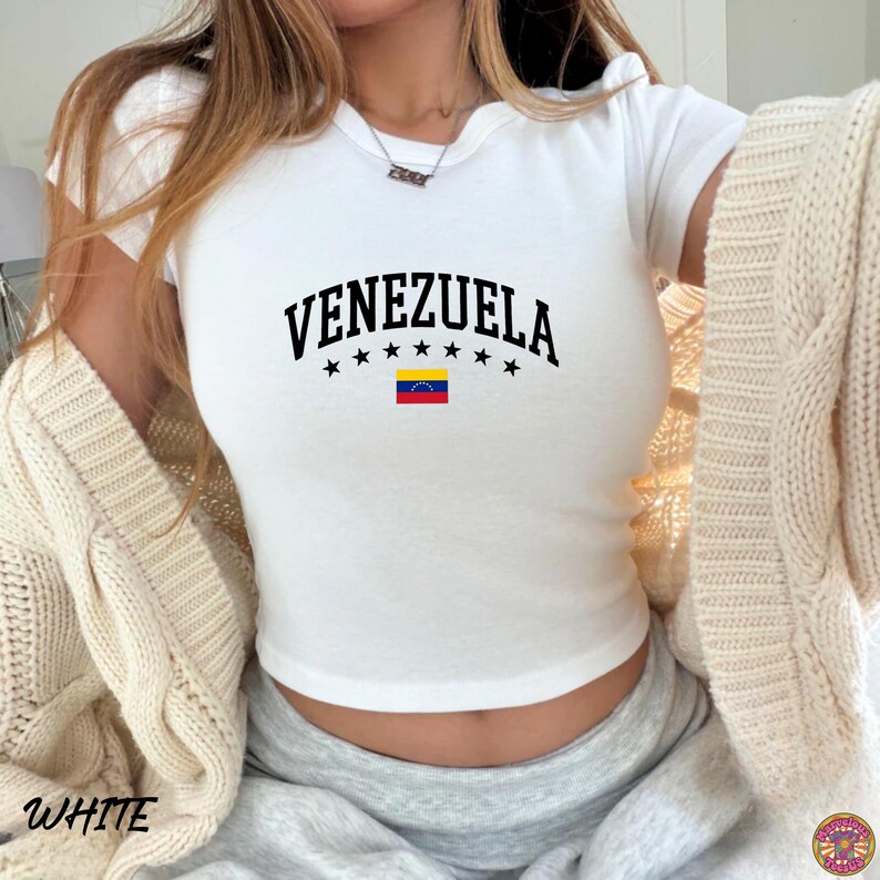 May include: White crop top with the word "VENEZUELA" in black, arched above a row of black stars and a small Venezuelan flag. The shirt is short-sleeved and has a scoop neck.