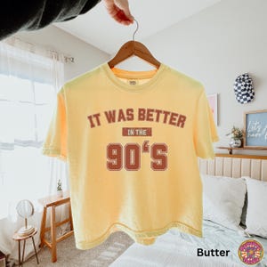 May include: A butter yellow short-sleeve t-shirt featuring the text IT WAS BETTER IN THE 90'S printed in a distressed dark red font. The shirt is displayed on a wooden hanger against a neutral background, showcasing a crew neckline and casual fit.