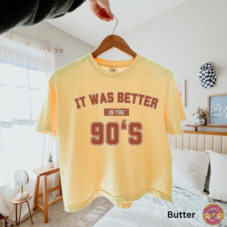 May include: A butter yellow short-sleeve t-shirt featuring the text IT WAS BETTER IN THE 90'S printed in a distressed dark red font. The shirt is displayed on a wooden hanger against a neutral background, showcasing a crew neckline and casual fit.