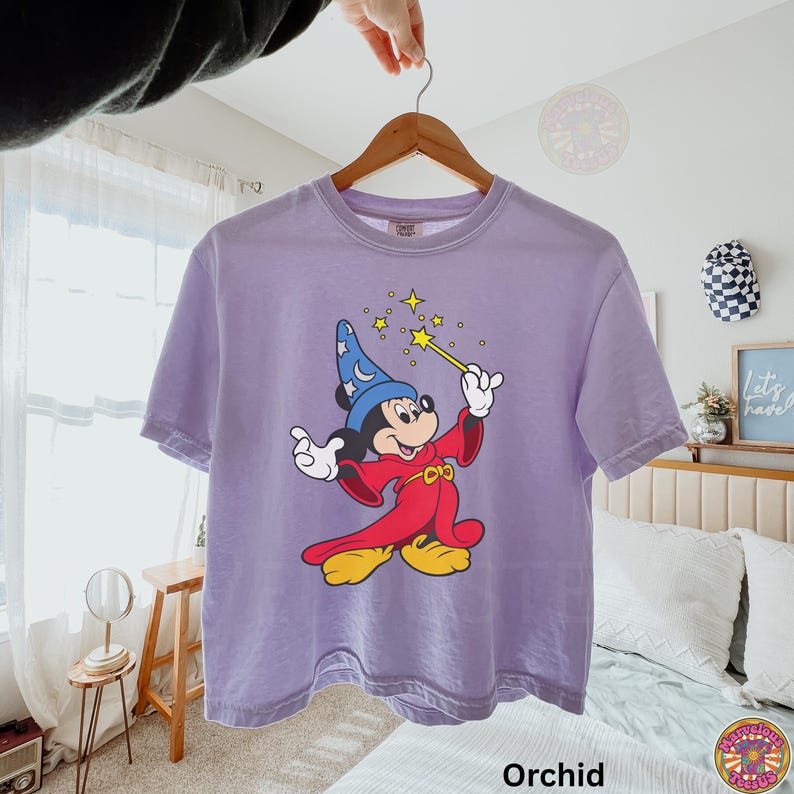 Puede incluir: Camiseta color orqu&iacute;dea con un gr&aacute;fico de Mickey Mouse vestido de mago, sosteniendo una varita con estrellas. El personaje de dibujos animados lleva un sombrero de mago azul y una t&uacute;nica roja. La camiseta est&aacute; colgada de una percha de madera.