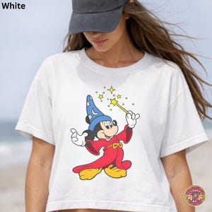 Puede incluir: Camiseta blanca con una ilustraci&oacute;n de dibujos animados de Mickey Mouse como mago, sosteniendo una varita con estrellas. El texto "White" est&aacute; en la esquina superior izquierda. El dise&ntilde;o incluye rojo, azul, amarillo y negro.