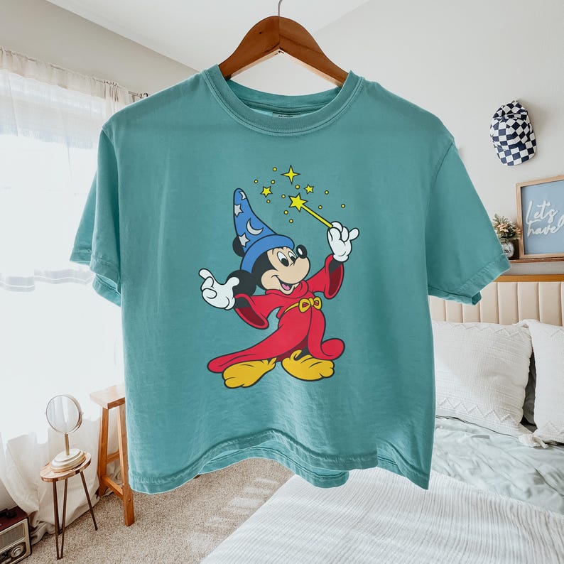 Puede incluir: Camiseta de color turquesa con un dibujo de Mickey Mouse como hechicero, sosteniendo una varita con estrellas. El personaje de dibujos animados lleva un sombrero azul, una t&uacute;nica roja y zapatos amarillos. La camiseta est&aacute; colgada de una percha de madera.