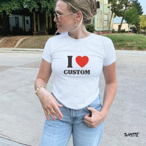 May include: White t-shirt with the text "I &hearts; CUSTOM" in black and red. The shirt is short-sleeved and worn with light blue jeans. The word "WHITE" is printed at the bottom right corner of the image.