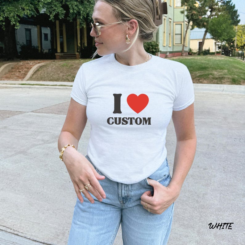 May include: White t-shirt with the text "I &hearts; CUSTOM" in black and red. The shirt is short-sleeved and worn with light blue jeans. The word "WHITE" is printed at the bottom right corner of the image.