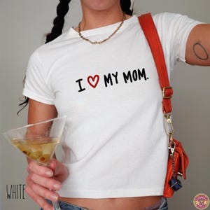 May include: White t-shirt with the text "I ♥ MY MOM" in black and a red heart. The shirt is paired with a red purse and a gold chain necklace. A martini glass with olives is held in the hand.