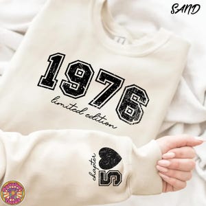 May include: Sand-colored sweatshirt with the year "1976" in black, distressed lettering. The words "limited edition" are below the numbers. The sleeve features a black heart and the number "5" with the word "chapter".