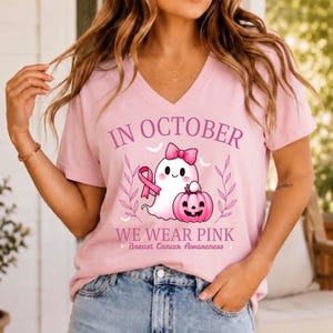 May include: A light pink V-neck t-shirt featuring a graphic of a white ghost with a pink bow holding a pink pumpkin and a pink ribbon. The text reads IN OCTOBER WE WEAR PINK Breast Cancer Awareness in purple and pink lettering.