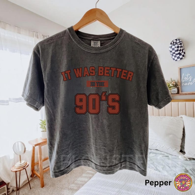 May include: A charcoal gray short-sleeve t-shirt with a vintage-style wash. The front features the text "IT WAS BETTER IN THE 90'S" printed in a bold, dark red font with a black outline, centered on the chest area of the garment.