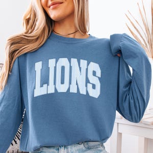 May include: A blue long-sleeve crewneck sweatshirt featuring the word LIONS printed in a white and blue vertical pinstripe font across the chest. The garment has a classic fit with ribbed cuffs and a ribbed neckline, showcasing a simple design.