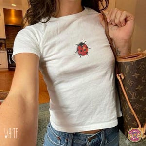 May include: A white short-sleeved t-shirt featuring a small, centered graphic of a red ladybug with black spots. The shirt has a crew neckline and a fitted silhouette, showcasing a simple design with a single insect illustration on the chest.