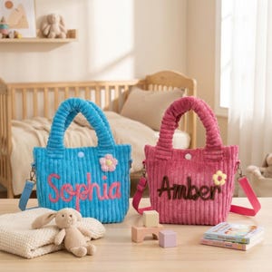 May include: Two corduroy tote bags, one turquoise with "Sophia" in pink, the other pink with "Amber" in brown. Each has a flower accent and a shoulder strap. The bags are on a wooden surface with children's items.