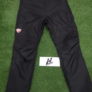 May include: Black motorcycle pants with reinforced panels and zippered pockets. Features a red and white Ducati logo patch on the left thigh. The pants have a button and zipper closure at the waist. The brand name "HOSIMAX" is visible on a white label.