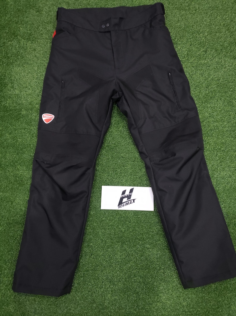 May include: Black motorcycle pants with reinforced panels and zippered pockets. Features a red and white Ducati logo patch on the left thigh. The pants have a button and zipper closure at the waist. The brand name "HOSIMAX" is visible on a white label.