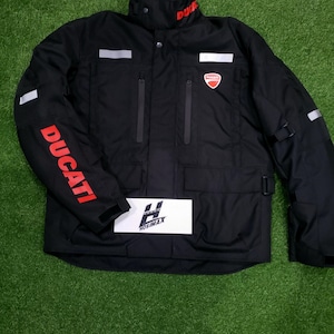 May include: Black Ducati motorcycle jacket with red Ducati lettering on the sleeve and collar. The jacket has a red Ducati logo on the chest, multiple pockets, and reflective stripes. The jacket is on a green surface.