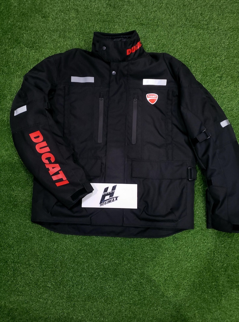 May include: Black Ducati motorcycle jacket with red Ducati lettering on the sleeve and collar. The jacket has a red Ducati logo on the chest, multiple pockets, and reflective stripes. The jacket is on a green surface.