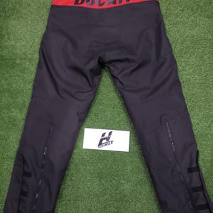 May include: Black motorcycle pants with a red waistband featuring the white word "DUCATI". The pants have zippers on the lower legs and black straps. The pants are displayed on a green surface.