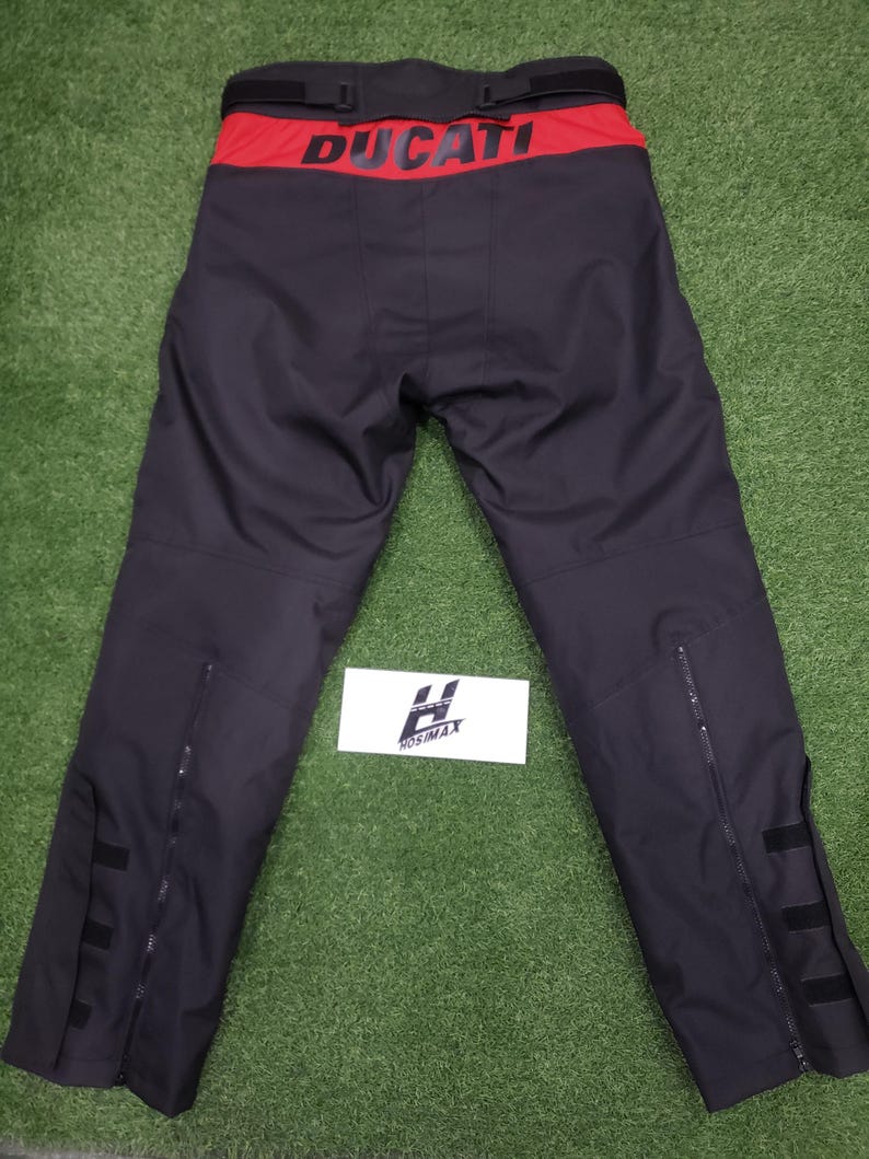May include: Black motorcycle pants with a red waistband featuring the white word "DUCATI". The pants have zippers on the lower legs and black straps. The pants are displayed on a green surface.