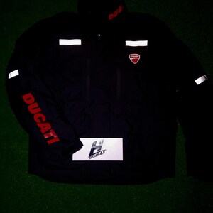 May include: Black Ducati motorcycle jacket with red Ducati lettering on the sleeve and collar. The jacket has reflective white stripes and the Ducati logo on the chest. The jacket also has a white rectangle with the text "HOSIMAX".