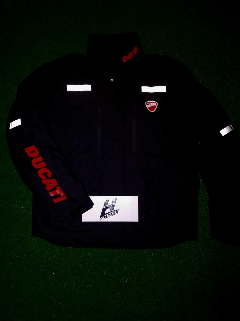 May include: Black Ducati motorcycle jacket with red Ducati lettering on the sleeve and collar. The jacket has reflective white stripes and the Ducati logo on the chest. The jacket also has a white rectangle with the text "HOSIMAX".